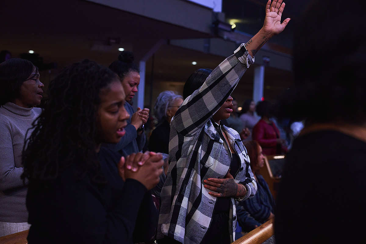 Woman lifting her hand in prayer and worship, surrounded by other congregants in deep spiritual connection.